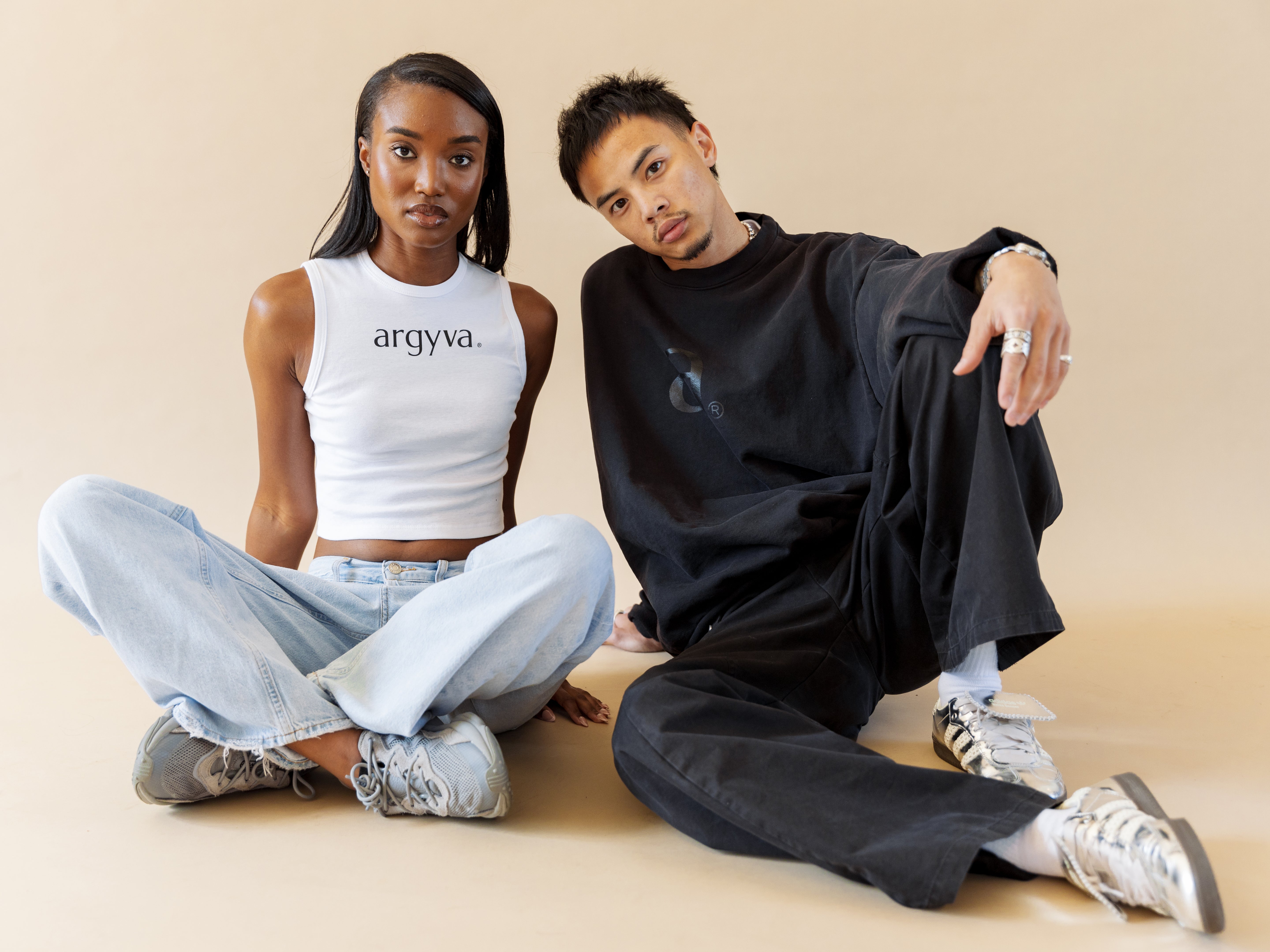 Two people sitting on the floor against a beige background, wearing 'argyva' clothing.